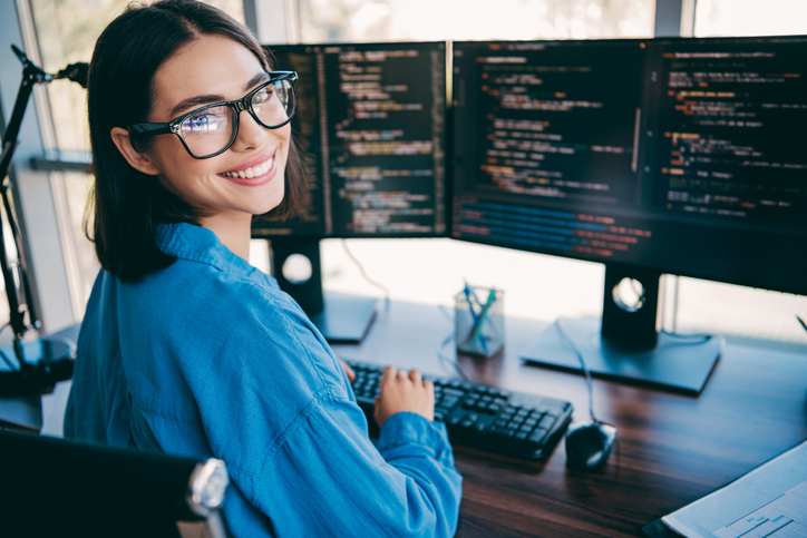 Desarrolladora web sonriendo frente a dos monitores con código, representando el trabajo de programación y desarrollo web profesional